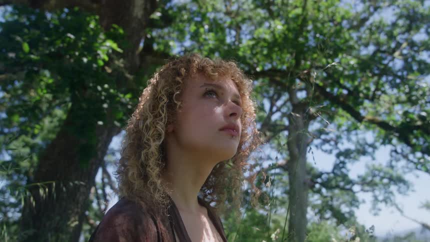 A woman with curly hair looks up at the sky among green trees
