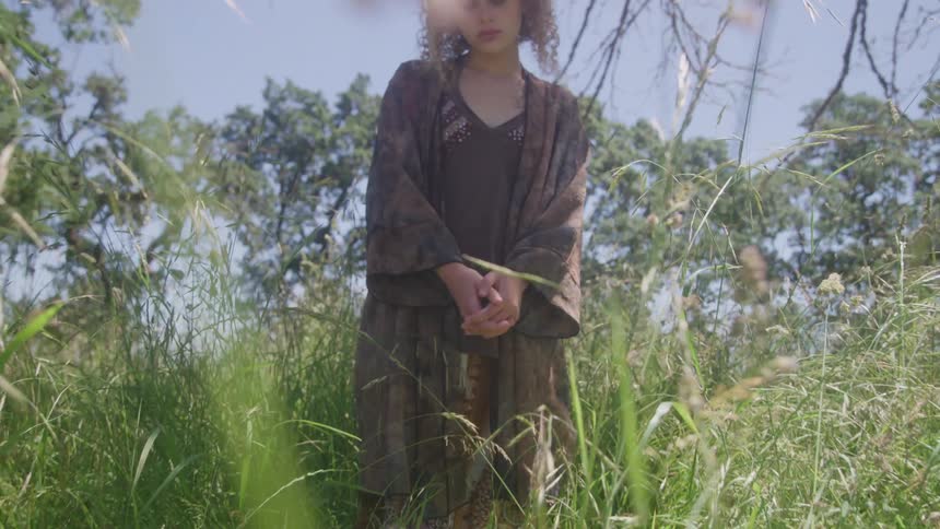 Woman with bowed head stands among tall grass under the sun