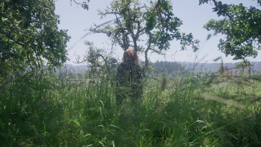 A young woman stands in tall grass beneath oak trees gazing toward distant hills on a bright calm day