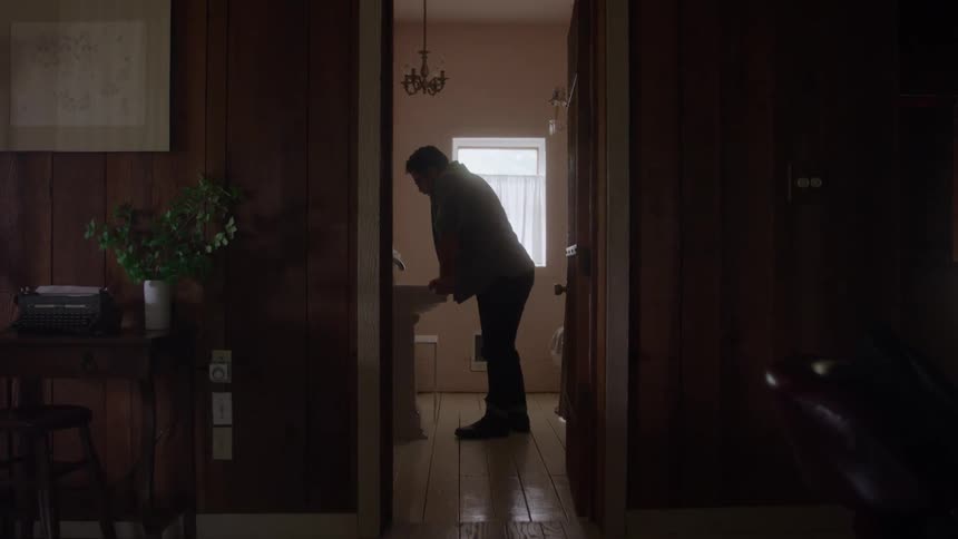 A person bends over a sink in a dimly lit hallway to a bathroom