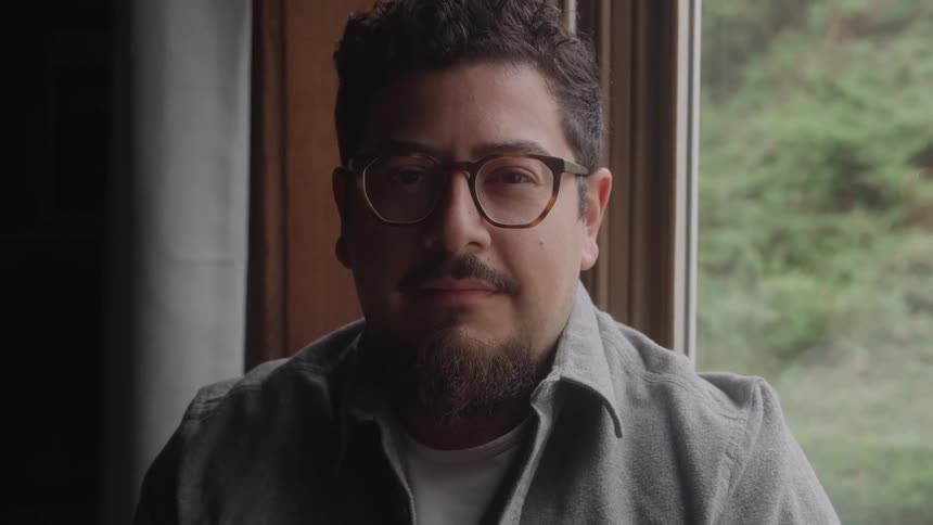 Man with beard and glasses smiles softly near the bright window