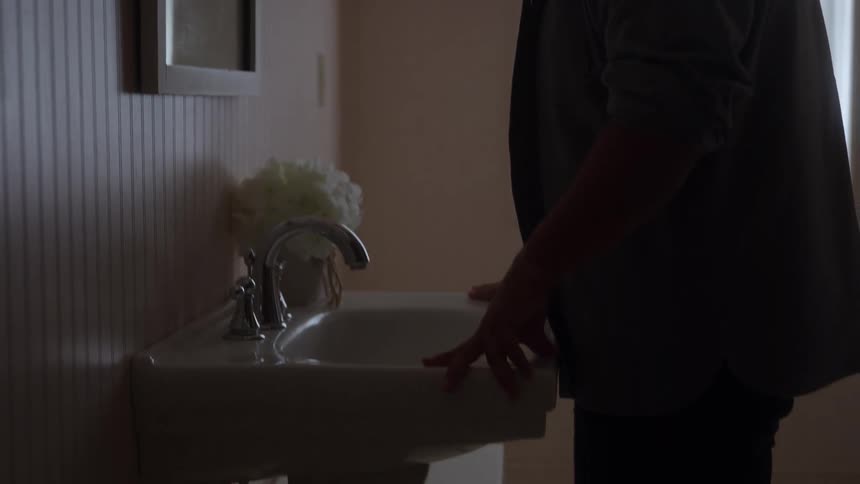 A person leans over a white wall mounted sink in a dimly lit bathroom