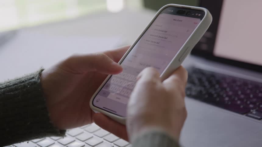 The video is a closeup shot of a persons hands holding a white smartphone and scrolling through the calendar application the hands are positioned over a laptop keyboard suggesting the person is working or managing their schedule on multiple devices