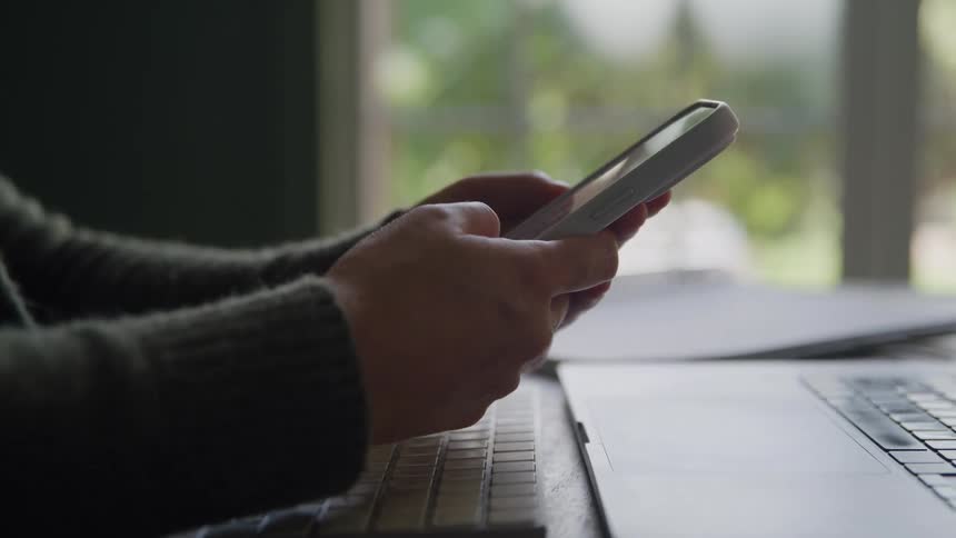 The video is a closeup shot of a persons hands holding and interacting with a white smartphone positioned next to an open laptop the scene is dimly lit indoors near a window with a blurry green background suggesting working from home or an office