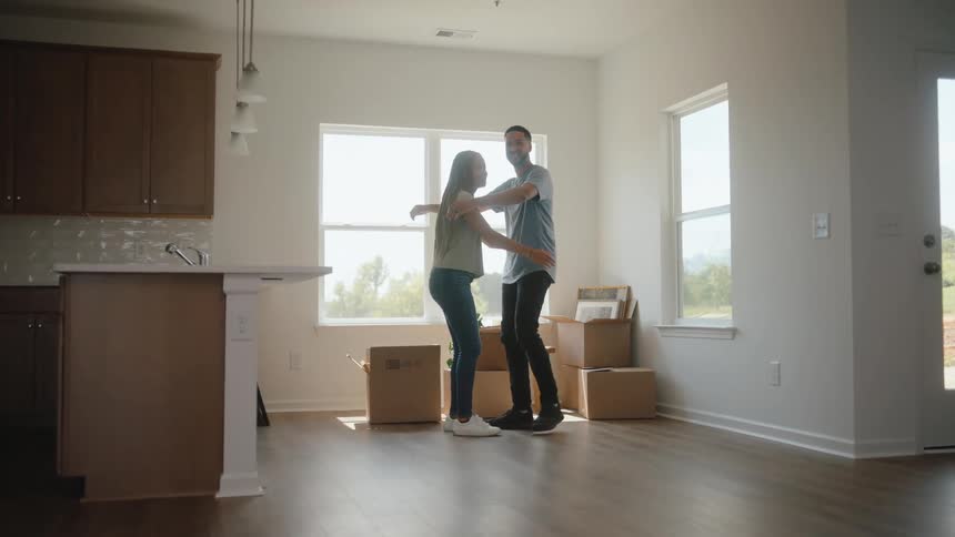 A man in a blue shirt and a woman in jeans hug and smile at each other in the center of an empty new house with hardwood floors large bright windows and moving boxes nearby