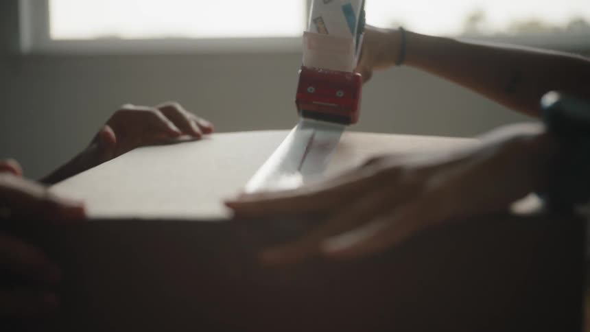 A black female smiles as she tapes up a box while a person across from her holds the box