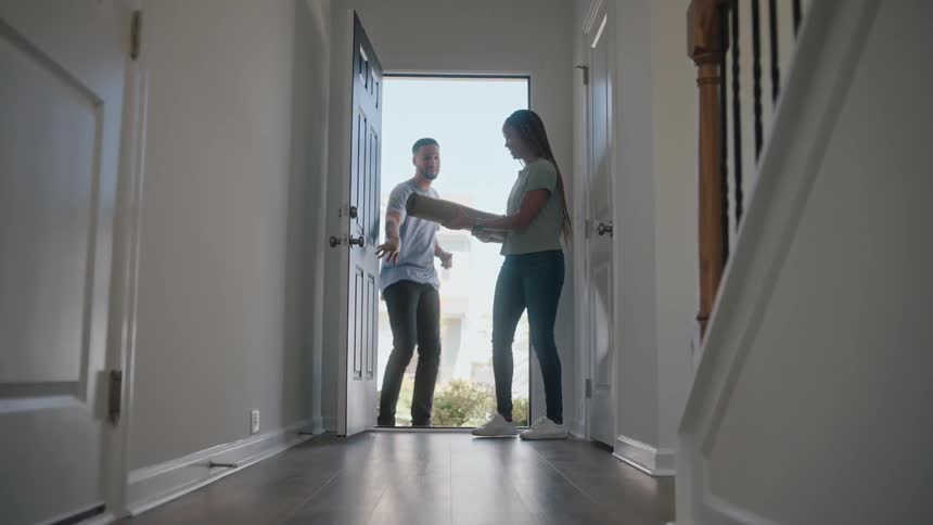 The video is a lowangle wide shot of a modern homes entryway with a smiling young woman opening the white front door and stepping inside a man is visible outside the door and then follows her in suggesting a couple is arriving home to a brigh