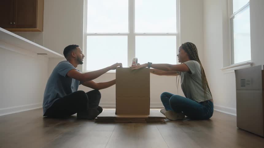 The video shows a young couple sitting on the floor of a bright empty room facing each other while sealing a large cardboard moving box the room has a large window letting in bright light and the scene suggests they are working together to pack or unp