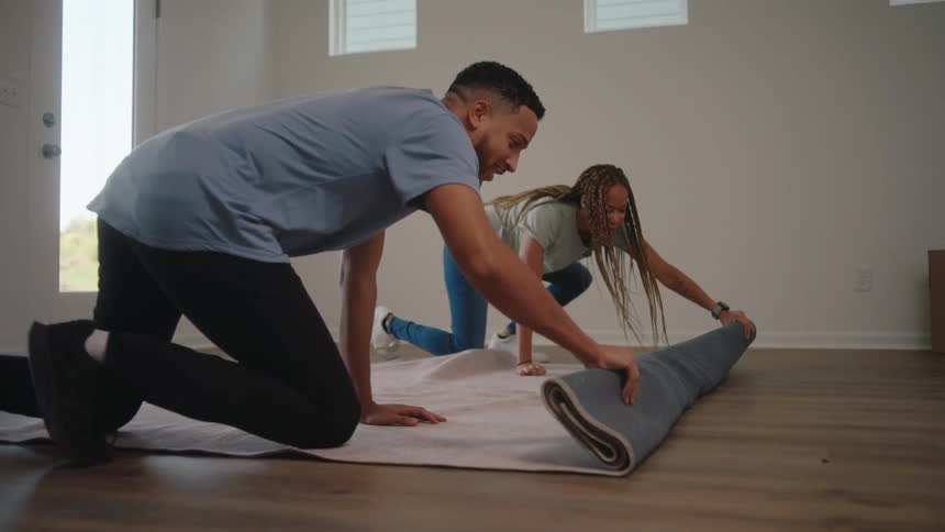 A smiling young couple unfolds a rug on the floor inside their cozy living room sharing a cheerful moment together