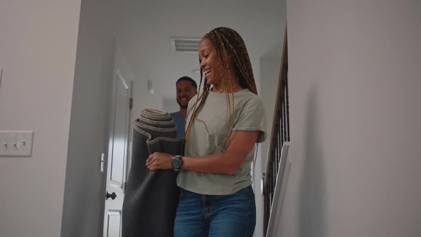 A smiling young couple carries a folded rug into their living room sharing a lighthearted moment as they prepare to lay it down together