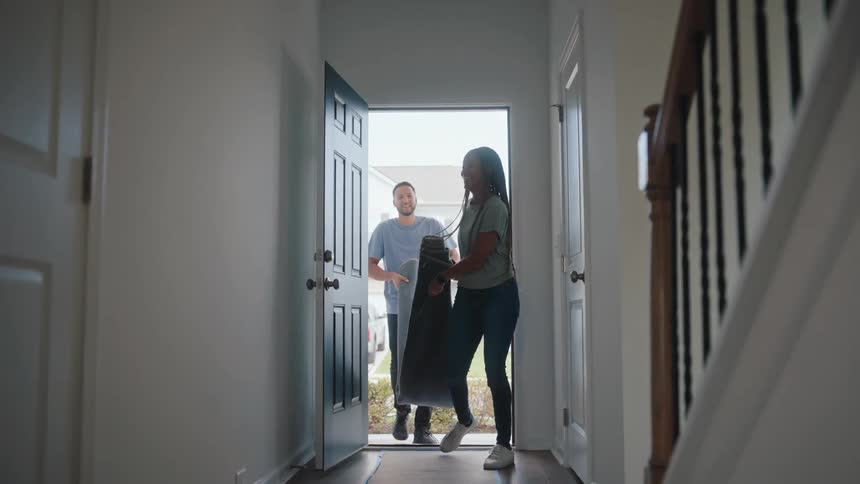 A smiling woman in jeans and a man in a light blue shirt carry a rolledup gray rug as they walk into the bright foyer of a new house through the open front door