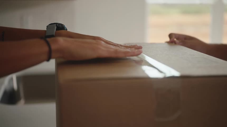 The video is a closeup shot of two peoples hands working together to seal a cardboard moving box with clear packing tape the scene is brightly lit by a large window in the background suggesting they are packing or moving into a new home