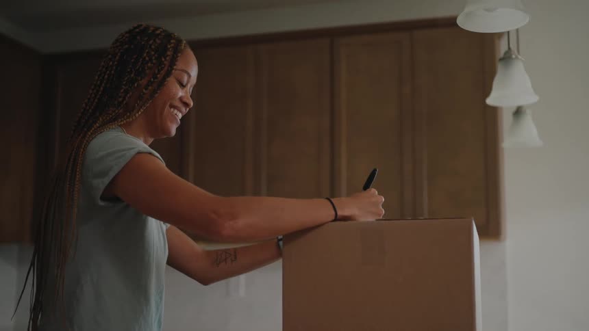 The video is a mediumcloseup shot of a young woman with long braids smiling as she signs a large cardboard moving box she is standing in a kitchen with dark wooden cabinets in the background suggesting she is packing or unpacking in a new home