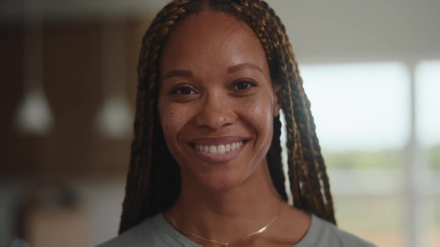 The video is a mediumcloseup shot of a young woman with long goldenbrown braids smiling and then looking directly at the camera she is standing indoors in a brightly lit space with a blurred lightcolored background