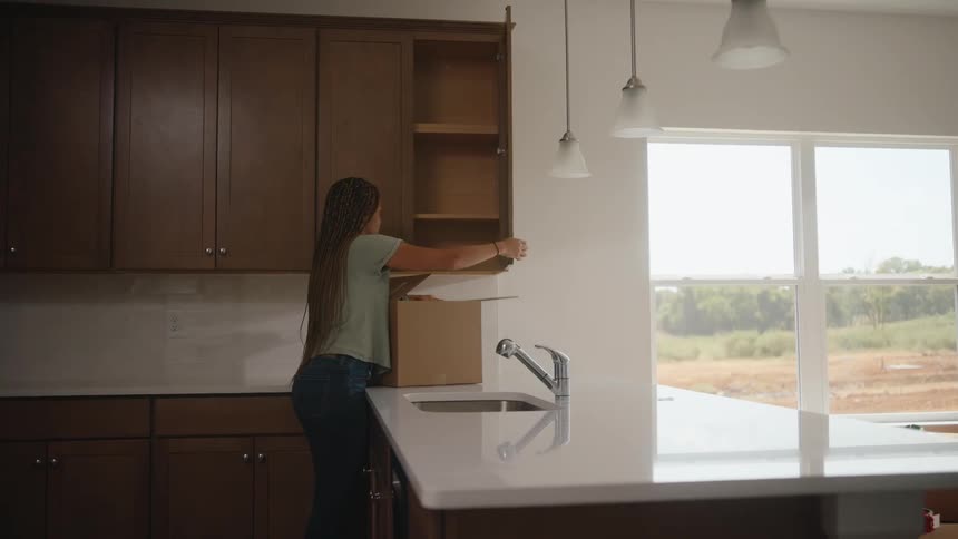 A young woman with long braids stands at a kitchen counter next to a box opening an upper wooden cabinet door and placing items from the box into the empty cabinet shelves in a kitchen with a large window