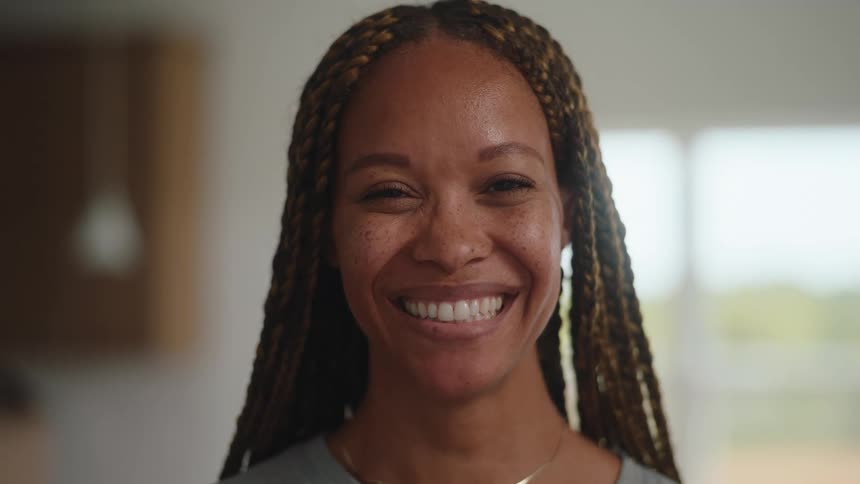 A smiling woman with long brown braids and freckles looks directly at the camera with a wide genuine smile standing in a bright modern indoor setting