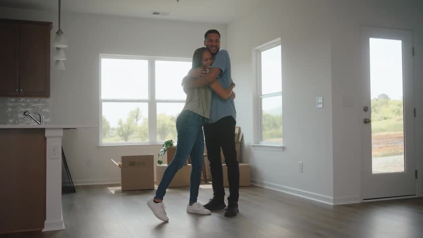 A woman in a green shirt and a man in a blue shirt embrace in the sunlit living area of their new home standing on hardwood flooring surrounded by moving boxes and kitchen cabinets