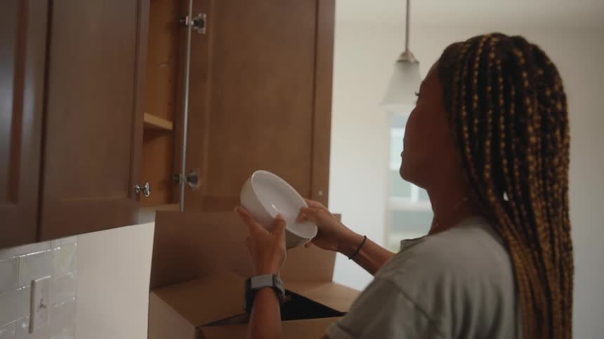 The video is a mediumcloseup shot of a black woman with long goldenbrown braids who is smiling faintly as she packs an open cardboard box the setting appears to be a brightly lit modern kitchen and the woman is busy with the task of moving into a n