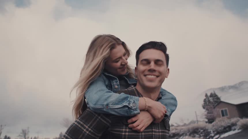 The video clip features a young man giving a young woman a piggyback ride outdoorsthey are both smiling broadly and appear happy with a snowy mountainous landscape and a few houses visible in the background under an overcast sky