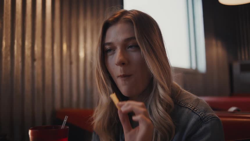 The video is a mediumcloseup shot of a young woman with long blonde hair and a denim jacket sitting in a dinerlike booth she is holding and looking at a french fry with a red cup and the restaurants slatted wall paneling visible in the background
