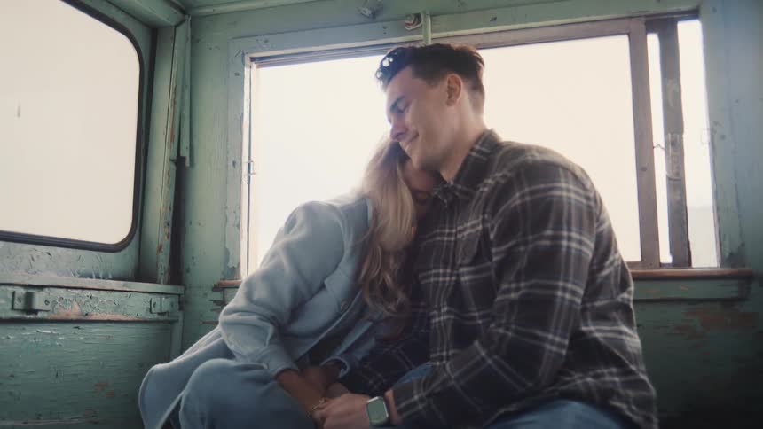A couple sits side by side inside a boat at midday smiling warmly as they hug each other under the sun