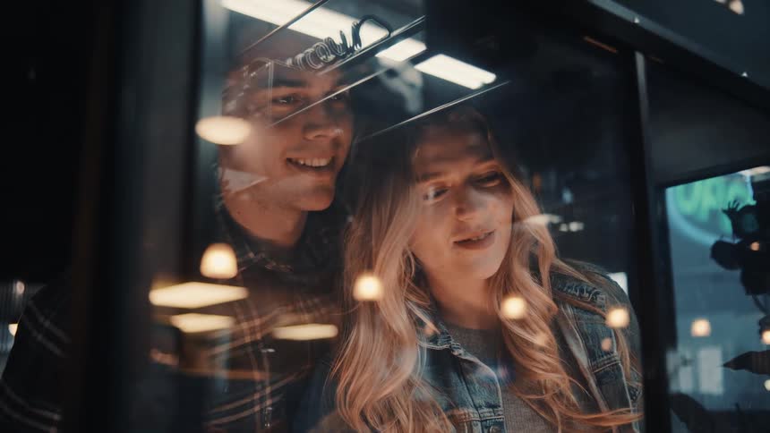 The video is a mediumcloseup shot of a young blonde woman and a man reflected in a cafe or store window they are both smiling and looking off to the side suggesting they are enjoying a conversation or observing something inside