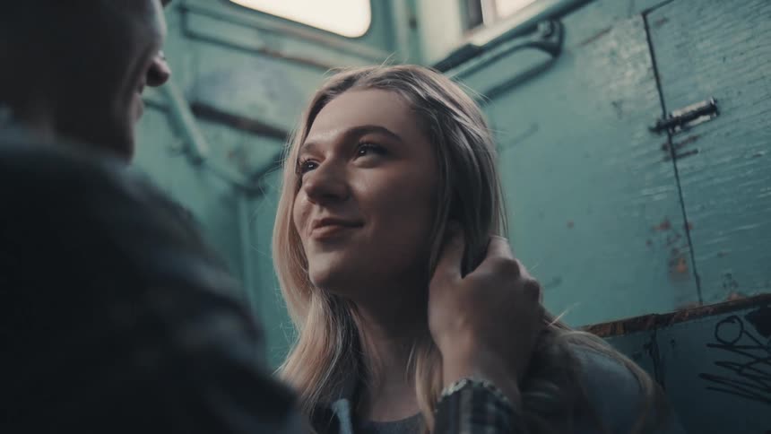 A woman with long light hair looks up smiling at an outoffocus manwhose hand is touching her cheek as the pair stand in a narrow space with blue graffiticovered walls