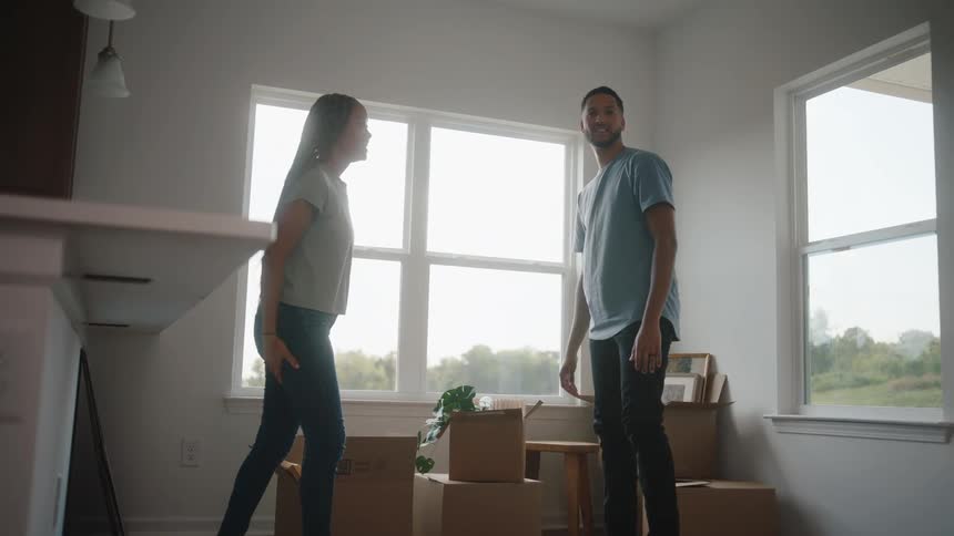 The video shows a young couple in a bright empty room with large windows squatting down as they place a potted plant into a cardboard box the woman has long blonde braids and the man wears a blue tshirt as they pack for a move or unpack in their new h