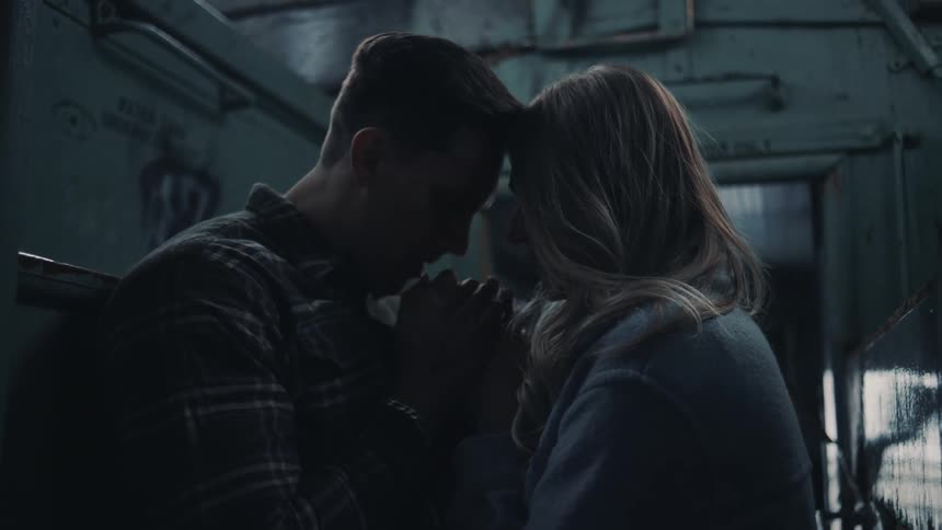 A man and a woman stand in a dark enclosed space with graffiticovered walls facing each other with foreheads almost touching while the man holds both of the womans hands near their chests