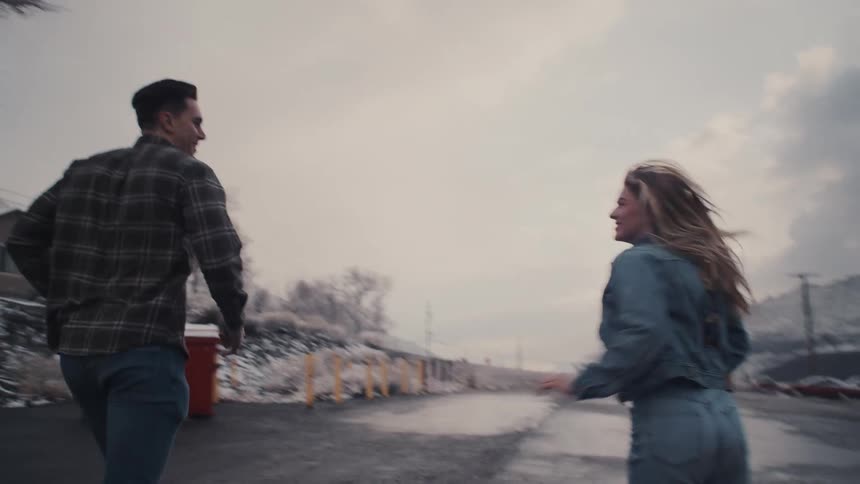 A young couple runs side by side playfully down the middle of a snowlined street at midday their faces lit with excitement and laughter