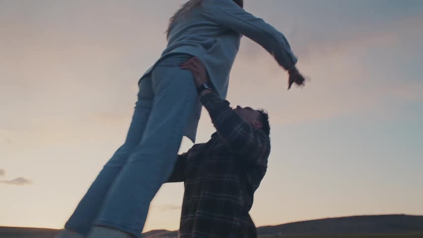 A man in a plaid shirt lifts a woman in light blue into the air at sunset over open fields capturing a playful joyful moment