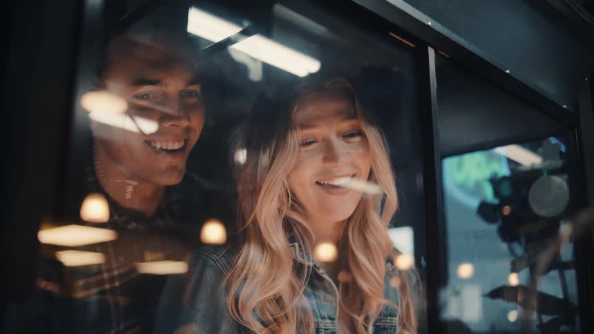 The video is a mediumcloseup shot of a young blonde woman with a denim jacket and a bright smile seen through a reflecting window or glass partition a man is also briefly visible behind her smiling and the reflections show bright busy lights from w