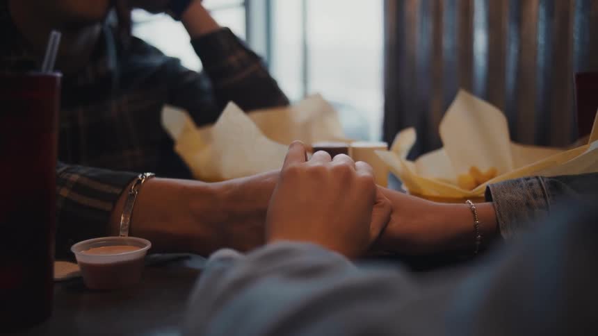 Two people sit at a table in a restaurant with hands resting together in the foreground while the man in a plaid shirt looks up and to the right with a slight smile and food wrapped in paper rests on the table