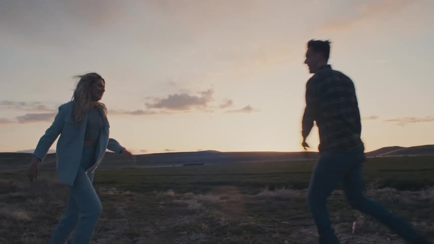 A man and a woman dance facing each other in a field with undulating terrain in the background under an evening sky