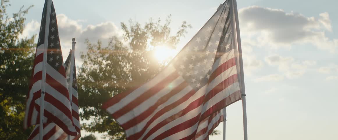 Multiple american flags are flying from poles and waving slightly against a bright sky and scattered clouds