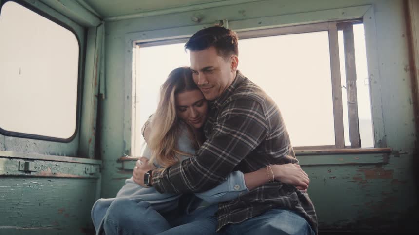 A man in a plaid shirt and a woman in a lightcolored hoodie are sitting facing forward and holding each other tightly in an embrace in a indoor setting with wooden walls and a large window behind them