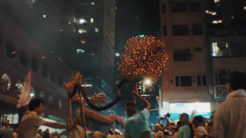 Multiple people carry a long dragon figure through a narrow city street at night with sparks and fire emanating from the objects head and body while tall buildings rise up in the background