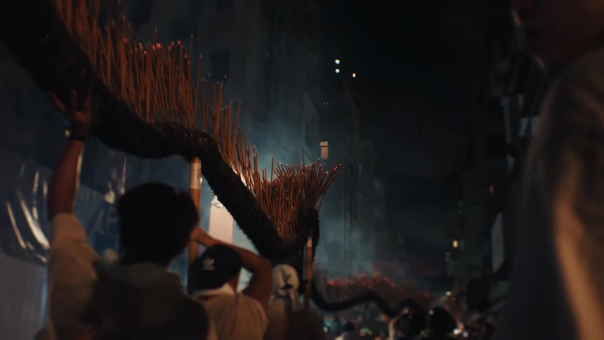 Multiple people carry a long dark curved object covered in burning incense sticks which emit sparks and light as they move through a crowded narrow city street at night with tall dark buildings in the background