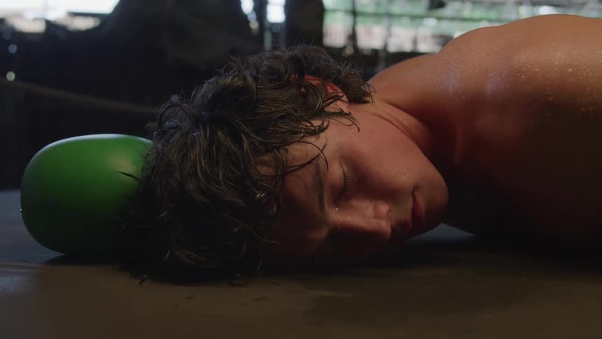 A young man lies facedown on the mat beside a green glove with eyes closed appearing dazed and exhausted