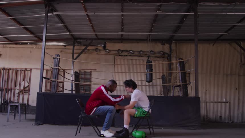 In a gritty gym a coach kneels by a seated boxer helping with wraps the scene feeling focused and supportive