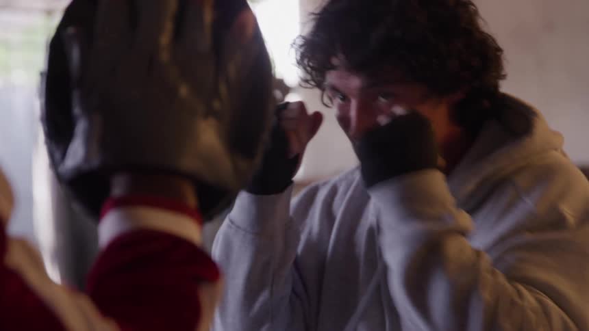A young male boxer in a gray hoodie punches a guard mitt someone is wearing eyes intense and determined