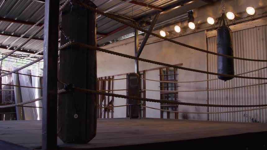 An empty boxing ring with wooden floor and thick ropes around the perimeter is shown in a rustic indoor space with a metal roof and a punching bag hanging in the background