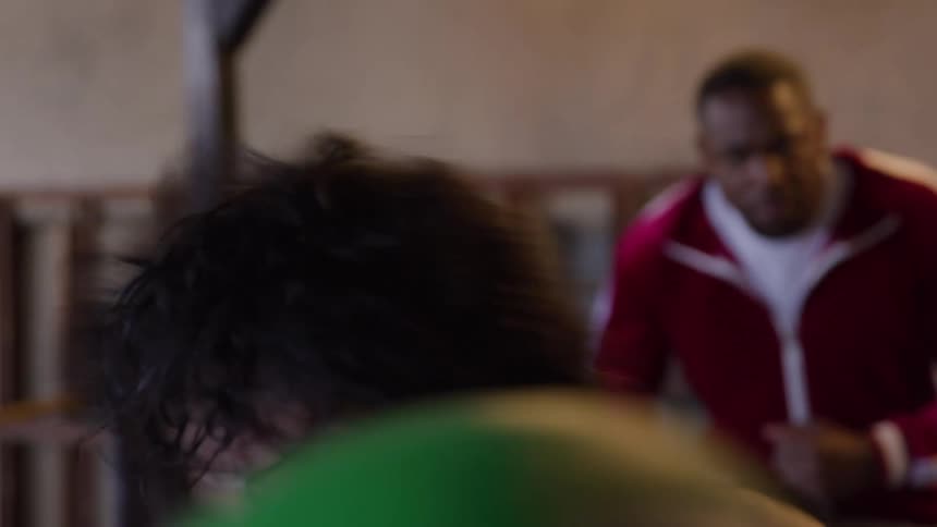 A boxer wearing green gloves in the foreground moves quickly while throwing punches toward the camera with a man in a red jacket acting as a trainer in the background