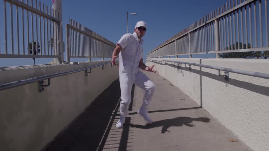 A person wearing all white clothing including a cap and sunglasses walks and dances along a concrete walkway between metal railings under a bright blue sky