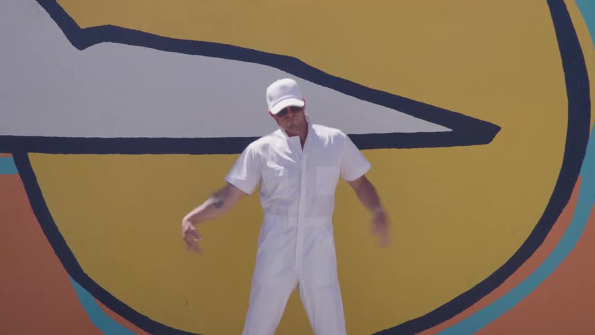 A man in an allwhite outfit dances in front of a bright yellow mural looking playful and confident