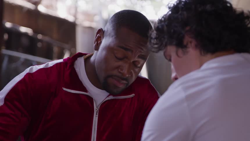 A man in a red zipup jacket leans in and speaks intently to a second person wearing a white shirt whose shoulder and back of the head are visible in the foreground in a dimly lit indoor space
