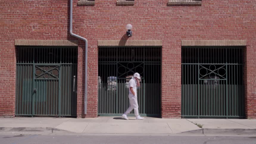Man dressed in white walks and dances in front of a brick building under the sunlight