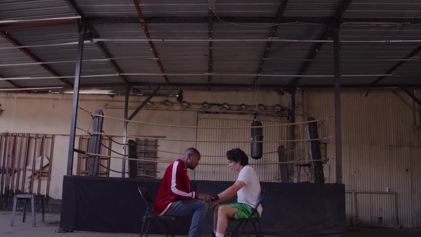 Male trainer conversing with a young male boxer while sitting indoors in front of a boxing ring during daytime