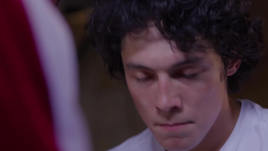A persons face with curly dark hair and light eyes looking intently toward an offcamera person whose red and white shoulder is visible in the foreground before glancing down and then back up again