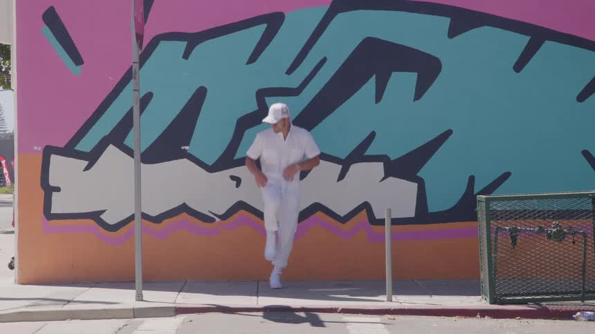 A person wearing all white clothing including a cap and sunglasses performs a dancing movement in front of a wall covered with bright teal and black graffiti on an urban sidewalk